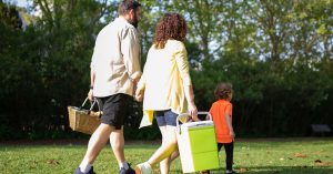 A family with a child carrying picnic essentials walking in a sunny park.