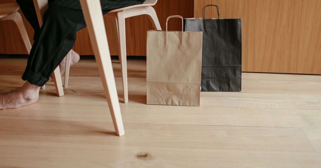 Side view of crop unrecognizable barefooted person resting on chair at table near paper bags placed on wooden floor at home