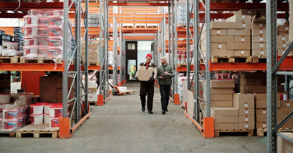 Two workers handle a package in a spacious warehouse surrounded by shelves stocked with boxes and products.