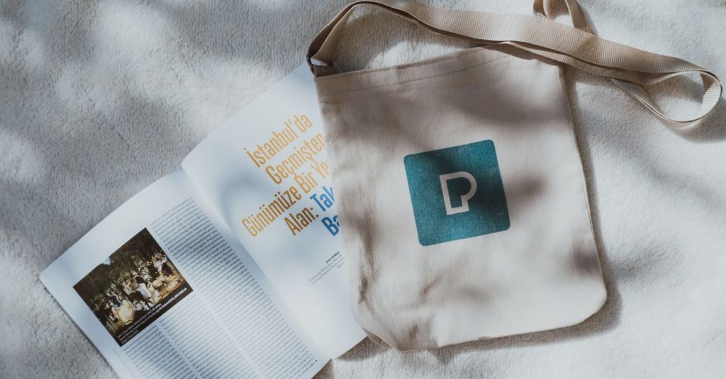 Linen tote bag with logo and open magazine on soft surface in natural light.