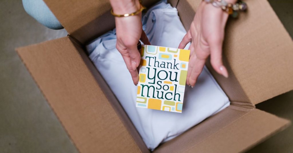 A person holds a thank you card above items in an open shipping box, suggesting care in sending.